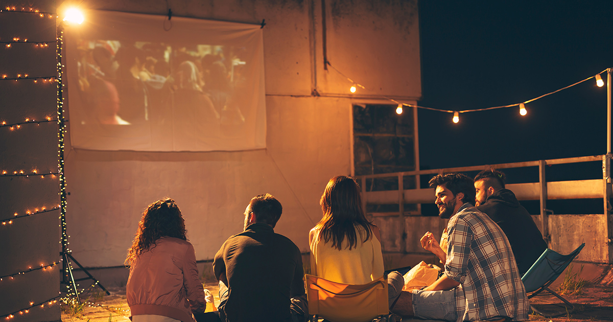 5 personer som sitter utomhus och myser med strandstolar, projektor visas på väggen och de tittar på film.
